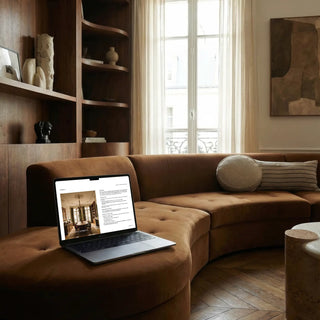 Laptop on a brown sofa in a stylish living room with a bookshelf and window in the background. The laptop screen shows an investment page.